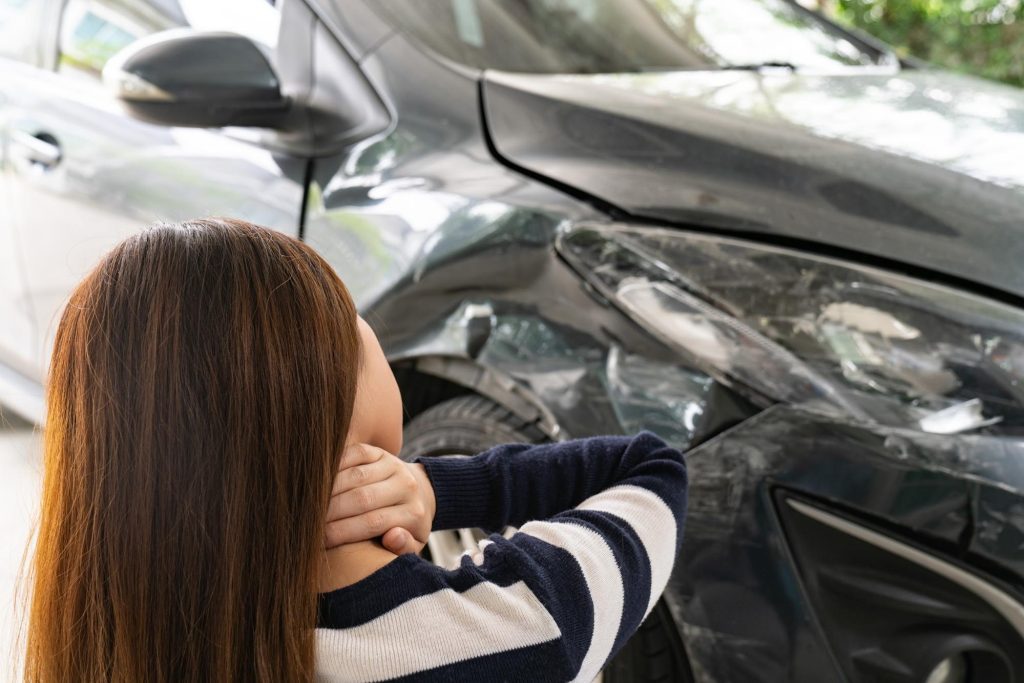 Woman with dark hair standing in front  of crashed car grasping neck while she appears to be in pain.