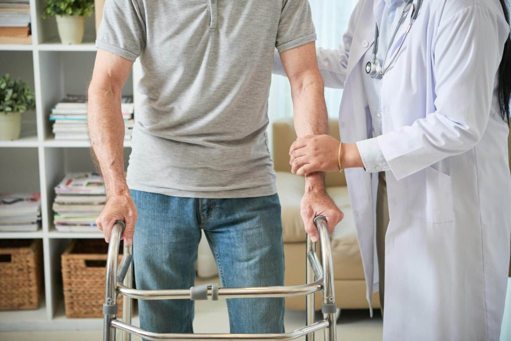 Young man at rehabilitation center, walking with walker while female doctor helps hold him up.