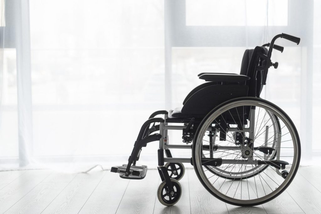 an empty wheelchair on a wood floor in front of a brightly lit window with sheer white curtains