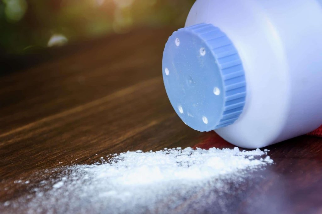 A close up photo of a bottle of talcum powder sitting on it's side on a wooden desk with a small pile of spilled talcum powder in front of the bottle.