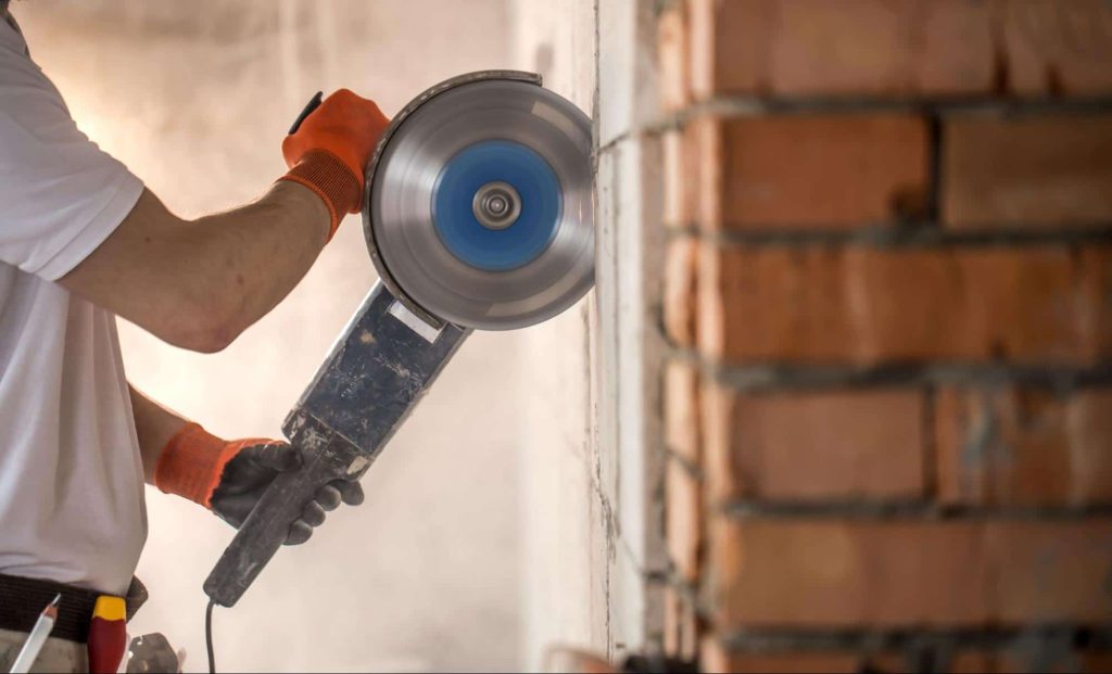 A construction worker uses an angle grinder to cut the mortar in a brick wall. Some jobs require working with dangerous tools. If a workplace injury happens, the workers are covered by workers compensation.