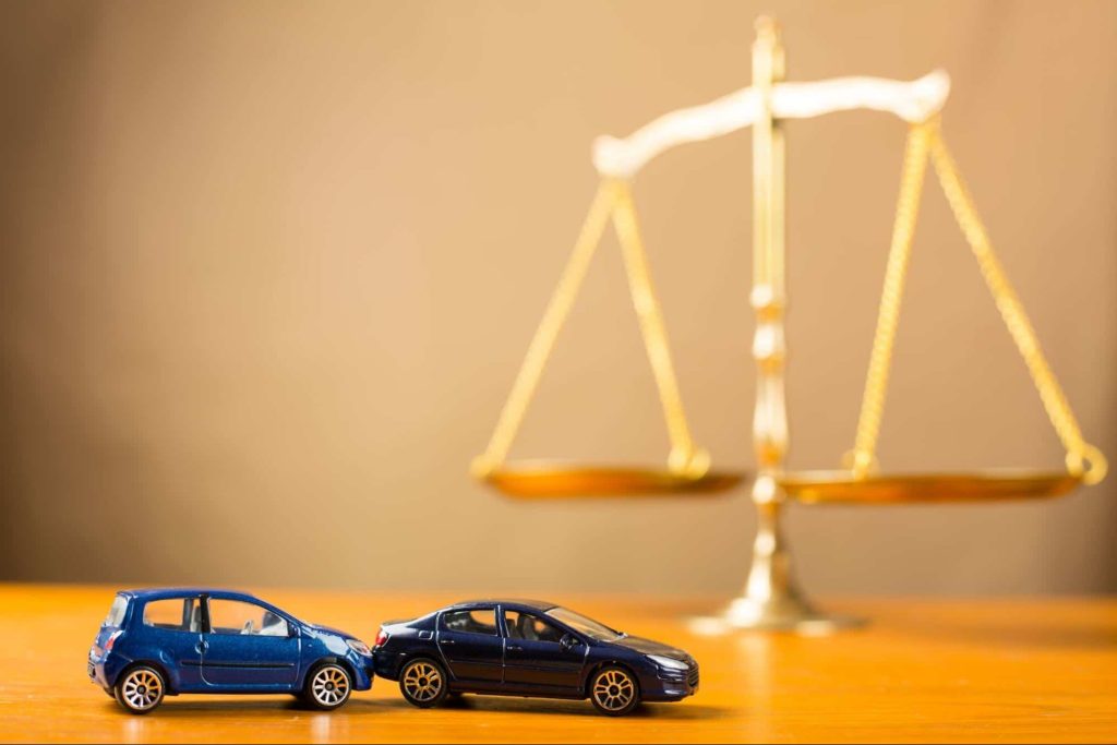 Toy cars on a wooden table arranged to look like a rear end car accident with the scales of justice in the background.